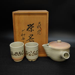 以下が含まれることがあります： 木箱、2つの陶器のカップ、急須がセットになった日本のティーセット。カップと急須はクリーム色で、カップと急須の蓋にはピンク色が入っています。木箱には日本語の文字が書かれています。