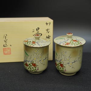 Puede incluir: Dos tazas de té de cerámica con tapas, con un diseño floral con detalles en rojo, blanco y verde. Las tazas son de color verde claro con un borde dorado y están colocadas frente a una caja de madera con texto japonés.