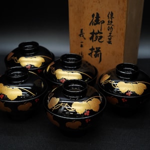 以下が含まれることがあります： 黒漆塗りの蓋付き椀5個セット。各椀には金箔と赤い実のデザインが施されています。背景には日本語の文字が書かれた木箱があります。椀は暗い表面に配置されています。