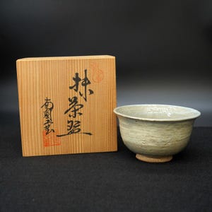 以下が含まれることがあります： 薄緑色の釉薬が施された陶器の茶碗が木箱の隣に置かれています。この茶碗は、表面にテクスチャがあり、小さく円形の台座が付いています。木箱には日本語の文字と赤いスタンプが押されています。