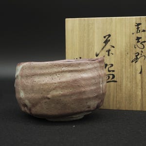 Op de afbeelding: Een getextureerde keramische theekom met een gespikkeld roze en beige glazuur, voorzien van een onregelmatige, met de hand gevormde rand. Achter de kom staat een lichtgekleurde houten doos met zwarte Japanse kalligrafietekens verticaal op de voorzijde.