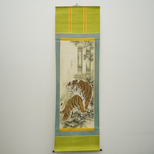 May include: A Japanese scroll painting featuring a tiger with brown and black stripes, set against a backdrop of bamboo trees. The artwork is framed by a green and blue border with yellow accents. The scroll is hanging on a white wall.