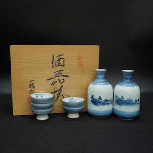 May include: A Japanese sake set featuring two blue and white ceramic bottles with landscape designs, small matching cups, and a wooden box with Japanese text and a red stamp.
