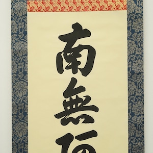 May include: A traditional Japanese scroll featuring black calligraphy on a cream-colored background. The scroll is framed with blue floral patterned fabric and a red and gold patterned border. The calligraphy is the focal point.