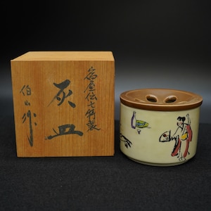 以下が含まれることがあります： 茶色の蓋と木箱が付いたベージュのセラミック容器。容器には人や物のイラストが描かれています。木箱には日本語の文字が書かれています。お香など用かもしれません。