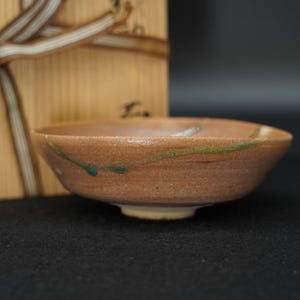 以下が含まれることがあります： 薄い緑色の釉薬の線と小さな青いアクセントが施された、質感のあるアースブラウンの浅い丸い陶器のボウル。ボウルは、装飾的な茶色のリボンで結ばれた明るい色の木箱の前に置かれています。