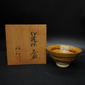 Puede incluir: Cuenco de cerámica artesanal con un diseño texturizado único en tonos marrones y amarillos. El cuenco se apoya sobre una base blanca. Junto a él, una caja de madera con caracteres japoneses.
