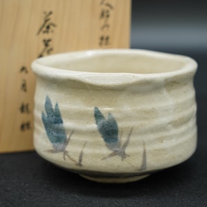 以下が含まれることがあります： ひび割れた釉薬仕上げのクリーム色の陶器製茶碗。外側には手描きの青い花のモチーフが施されています。茶碗はわずかに波打った質感の表面をしており、黒い日本語の書が書かれた木箱の前に置かれています。