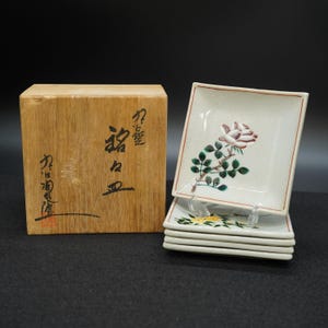 以下が含まれることがあります： 木箱の隣に積み重ねられた、花の模様が施された正方形のセラミックプレートのセット。プレートには、白、緑、黄色のバラやその他の花が描かれています。箱には日本語の文字が書かれています。