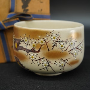 May include: A cream-colored ceramic tea bowl featuring a hand-painted design of brown branches with white plum blossoms and yellow centers. The bowl has a smooth, glazed finish and is positioned in front of a tan box tied with a blue ribbon.
