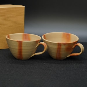 以下が含まれることがあります： オレンジ色の縦縞と曲がった取っ手が付いた、ベージュ色の陶器のティーカップ2つ。カップは暗い面に置かれ、背景には木製の箱があります。カップは素朴で手作りの外観をしています。