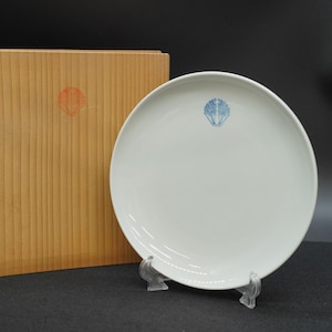 以下が含まれることがあります： 青と白のエンブレムが付いた白いセラミックプレート。透明なスタンドに展示されています。左側には赤いエンブレムが付いた木箱があります。プレートは丸く、表面は滑らかです。