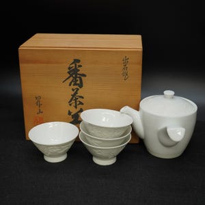 Könnte beinhalten: Ein weißes Keramik-Teeservice mit einer Teekanne, vier kleinen Schalen und einer Holzkiste. Die Teekanne und die Schalen haben ein Blumenmuster. Die Holzkiste hat japanische Schriftzeichen.
