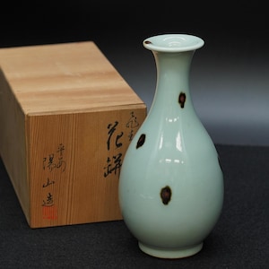 Può includere: Un vaso in ceramica verde pallido con macchie marrone scuro, accanto a una scatola di legno con calligrafia giapponese. Il vaso ha un collo sottile e un corpo arrotondato. La scatola di legno è marrone chiaro e ha un design semplice.