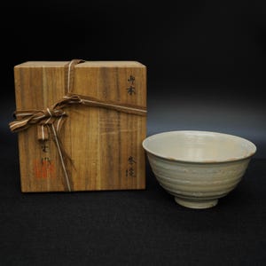 Peut inclure: Un bol en céramique de couleur claire avec un large bord et une base à pied est posé à côté d'une boîte en bois attachée avec un ruban marron. La boîte comporte des caractères japonais et un tampon rouge. Le bol a une surface texturée.