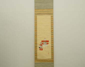 May include: A long, rectangular Japanese scroll painting. The top and bottom sections are a muted green with a patterned border. The central section is a light beige with a painted scene of figures in red and white. The scroll is suspended by a wooden hanger.