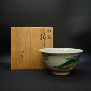 May include: A ceramic bowl with a cream-colored interior and exterior, featuring a painted landscape design in green, blue, and brown. The bowl sits next to a wooden box with Japanese calligraphy.