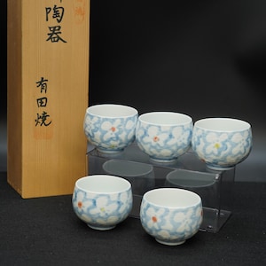 May include: Five Japanese ceramic tea cups with a floral design in blue, white, and red. The cups are on a clear acrylic stand, beside a wooden box with Japanese text. A traditional tea set.