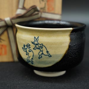 May include: A ceramic tea bowl featuring a two-tone design with a textured black exterior and a smooth cream-colored panel. The cream section displays a blue line drawing of two rabbits in motion. The interior of the bowl is finished in black.