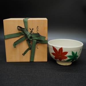 Puede incluir: Un cuenco de té de cerámica con diseños de hojas de arce rojas y verdes, junto a una caja de madera atada con una cinta verde. El cuenco tiene un interior blanco y una pequeña base. La caja tiene caracteres japoneses.