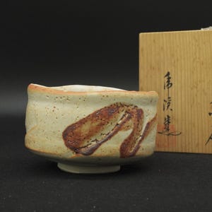 May include: A ceramic tea bowl with a cream-colored, speckled glaze and a prominent, dark brown, abstract brushstroke design. The bowl has an uneven, hand-formed rim and sits next to a light-colored wooden storage box featuring black Japanese calligraphy.
