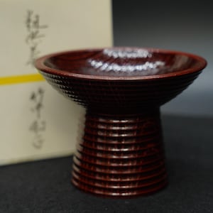 Puede incluir: Una copa de pedestal de madera de color marrón rojizo oscuro con un borde ancho y acampanado y una base cilíndrica. La superficie exterior está decorada con una serie de ranuras horizontales talladas que envuelven todo el cuerpo del recipiente.