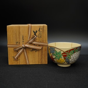 Könnte beinhalten: Cremefarbene Keramikschale mit einem Blumen- und Blattdesign in Rot, Grün, Blau und Gelb. Die Schale steht neben einer Holzkiste, die mit einem braunen Band gebunden ist. Die Kiste hat japanische Schriftzeichen.