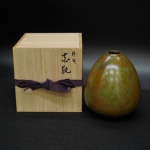 以下が含まれることがあります： 卵型の小さな花瓶と木箱。花瓶はブロンズ仕上げで、緑と茶色の斑点模様です。木箱は薄い色の木でできており、紫色のリボンで結ばれており、前面には日本語の文字が書かれています。