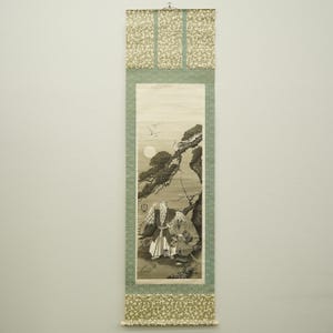 以下が含まれることがあります： 伝統的な衣装を着た2人の人物が木の下に座っている様子を描いた日本の巻物絵。背景には満月と鳥が描かれています。巻物には、幾何学模様の緑色の縁取りと、上部と下部に花のアクセントがあります。