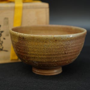 以下が含まれることがあります： 茶褐色の斑点模様がある丸い陶器製の茶碗。広い開口部と円形の高台が特徴です。外側には微細な横方向の筋があり、土の質感を活かした素朴な仕上がりになっています。全体的に落ち着いた色合いで、厚みのある縁が特徴的な形状です。