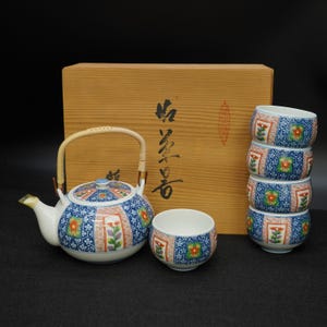 Puede incluir: Juego de té japonés con tetera, tazas y una caja de madera. La tetera y las tazas tienen un diseño floral azul y blanco con detalles en rojo y verde. La tetera tiene un asa trenzada.