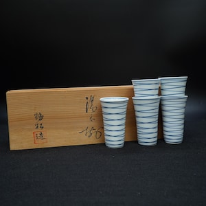 以下が含まれることがあります： 青い水平線が入った白い陶器のカップ6個セット。木箱の隣に展示されています。木箱には日本語の文字と赤いスタンプがあります。カップは円筒形で、ミニマリストなデザインです。