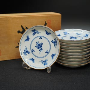 以下が含まれることがあります： 青い花柄とスカラップエッジが特徴の白い磁器皿のセット。 皿は茶色の縁があり、黒い表面に積み重ねられています。 背景には木製の箱があります。