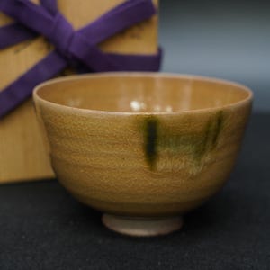 May include: A handcrafted, ceramic tea bowl with a textured, crackled surface in shades of tan and brown. The bowl features a small, circular foot and a dark green vertical accent. A wooden box with a purple ribbon is in the background.