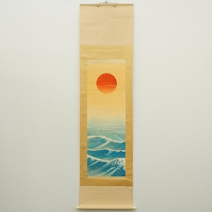 以下が含まれることがあります： 青い波の海に沈む鮮やかなオレンジ色の太陽を描いた、縦型の日本の巻物絵画。 このアートワークは、暖かく黄色がかったオレンジ色の空を特徴とし、ベージュの縁で縁取られています。