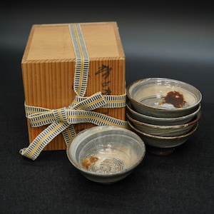 以下が含まれることがあります： 素朴なデザインのセラミックボウルのセット。縞模様のリボンで結ばれた木箱の隣に積み重ねられています。ボウルはグレーとベージュの色合いで、茶色のアクセントがあります。木箱には日本語のテキストがあります。