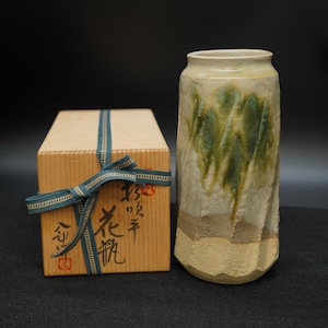 Könnte beinhalten: Eine handgefertigte Keramikvase mit strukturierter Oberfläche und grün-beiger Glasur. Daneben eine Holzbox, die mit einem blauen Band gebunden ist. Die Box hat japanische Schriftzeichen und einen roten Stempel.