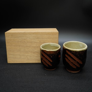 Puede incluir: Dos tazas de cerámica con base marrón oscuro y rayas diagonales naranjas. Los bordes son de color beige claro. Una caja de madera está en el fondo. Las tazas son probablemente para beber té o sake.