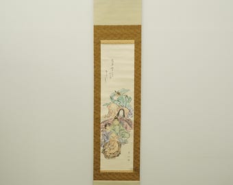 May include: A vertical Japanese scroll painting featuring a detailed illustration of figures in colorful robes. The artwork includes calligraphy and is framed by a textured brown border. The scroll is suspended from a wooden bar.