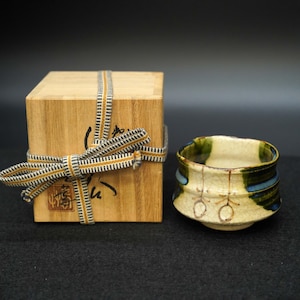 以下が含まれることがあります： クリーム色の外観に青と緑のアクセントが施された陶器のティーボウル。 ボウルの隣には、模様入りのリボンで結ばれた木箱があります。 箱には日本語の文字が書かれています。