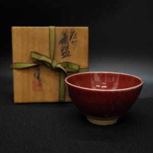 Può includere: Una ciotola in ceramica rosso scuro con bordo e base colorati chiari. Sullo sfondo una scatola di legno legata con un nastro verde. La scatola presenta caratteri giapponesi e un timbro rosso.