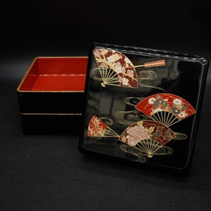 Op de afbeelding: Een zwarte gelakte doos met een rode binnenkant en een deksel versierd met gestileerde waaiers in rood, goud en wit. De doos is vierkant en heeft een glanzende afwerking, met traditionele Japanse designelementen.