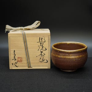 Puede incluir: Un cuenco de té de cerámica marrón con una superficie texturizada y un borde marrón claro. Al lado hay una caja de madera con caligrafía japonesa y una cinta decorativa. La caja está atada con una cinta a rayas beige y negra.