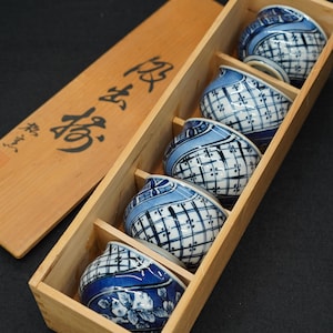 May include: A set of five blue and white ceramic cups with a geometric pattern, nestled in a wooden box. The cups have a rounded shape and a contrasting blue rim. The box has Japanese characters on the lid.