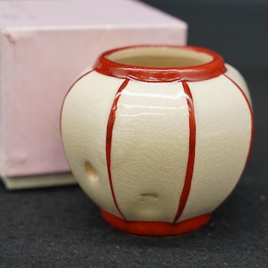 Może przedstawiać: Małe, okrągłe naczynie ceramiczne z kremowym szkliwem o spękanym wykończeniu. Przedmiot posiada pionowe, czerwone malowane paski biegnące od górnej krawędzi do podstawy oraz małe, okrągłe wgłębienie na boku zaokrąglonego korpusu naczynia.