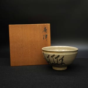 以下が含まれることがあります： ベージュ色の陶器のボウル。濃い茶色の筆致と小さな足が付いています。横には日本語の文字が書かれた木箱があります。ボウルはわずかにテクスチャ加工された表面と丸みを帯びた形状をしています。