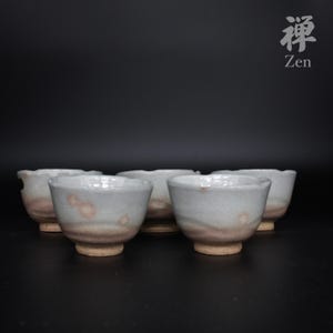 以下が含まれることがあります： 手作りの小さな陶器の器が5つ並んだ写真。淡いグレー色で、ピンクと茶色のアクセントがある。縁は少し波打ったデザイン。写真には「禅」の文字が筆文字で書かれている。