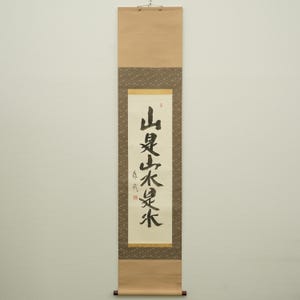 Peut inclure: Un rouleau japonais traditionnel avec calligraphie à l'encre noire. Le rouleau présente un fond beige avec une bordure à motifs et un rouleau en bois en bas. La calligraphie est verticale.