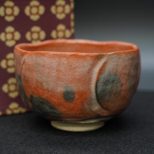 以下が含まれることがあります： 縁が波打った陶器製の茶碗。オレンジとくすんだ赤のひび割れた釉薬が施され、濃い灰色の円形の斑点がアクセントになっています。茶碗には、釉薬のかかっていない明るい色の円形の高台があり、外側は滑らかな曲線を描いています。