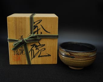 May include: A ceramic tea bowl with a dark interior and a textured exterior in shades of brown and tan. It sits next to a wooden box tied with a green ribbon and a dark green and brown striped ribbon. The box has Japanese characters.